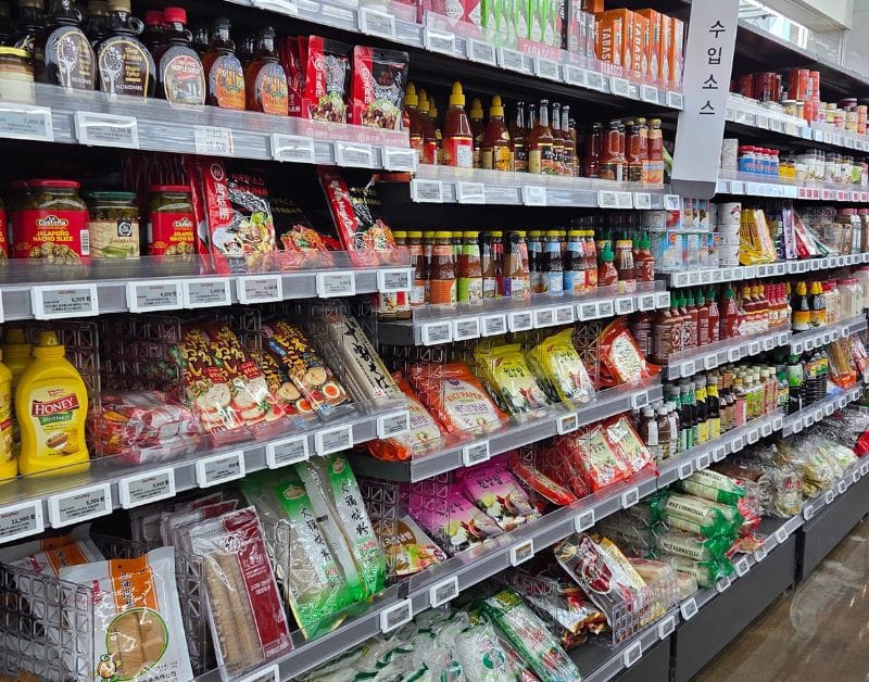 Supermarket shelf stocked with various imported sauces like Tabasco, Sriracha, and pasta sauce.