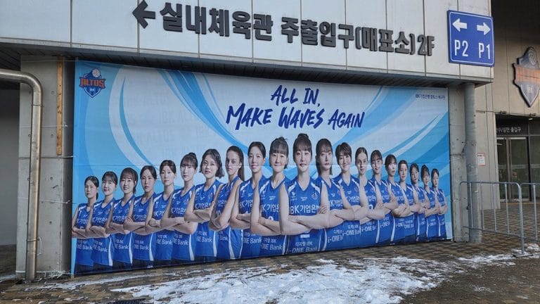 A large blue banner featuring the IBK Altos team players and the slogan "ALL IN, MAKE WAVES AGAIN" displayed outside the Hwaseong Indoor Arena before a Korean Volleyball Game.