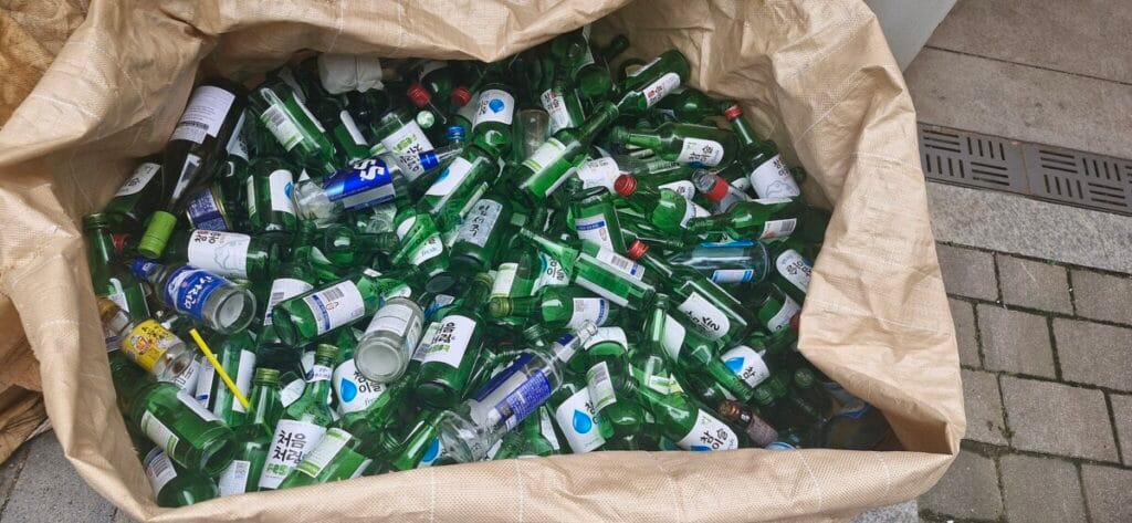 A large tan recycling bag filled to the brim with green and clear glass soju bottles outside a South Korean apartment.