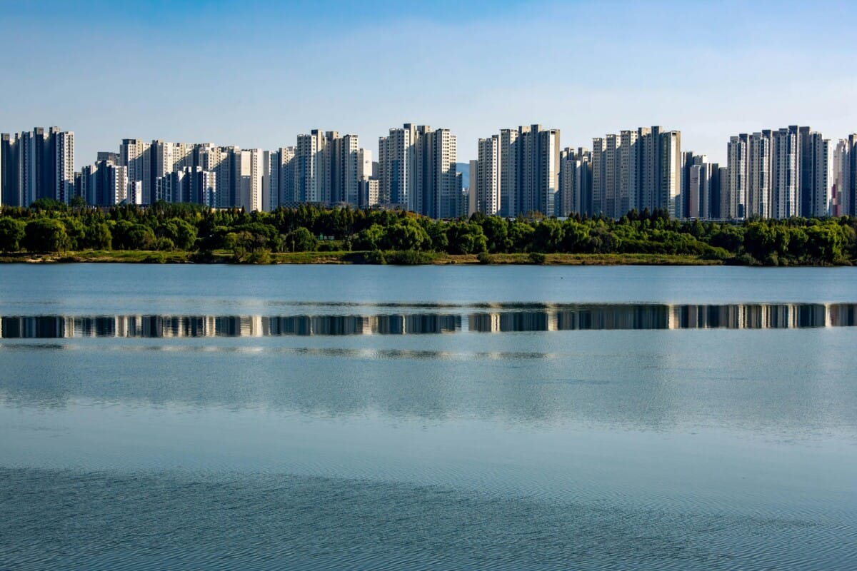 Modern high-rise apartment buildings in Korea reflected in a calm lake under a blue sky.