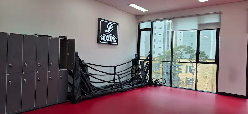 개인 사물함 위에 설치된 엘복싱 로고 간판과 벽쪽으로 접혀있는 검은색 복싱 링.