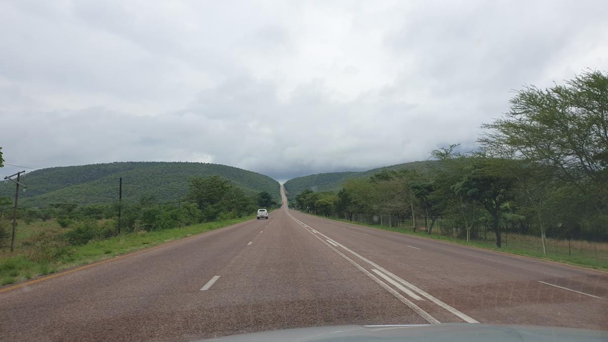 A vehicle drives along a paved road winding through a lush, hilly landscape under a blue sky.