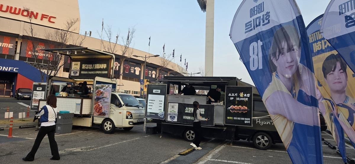 People stand near a red food truck parked next to a white food truck outdoors.