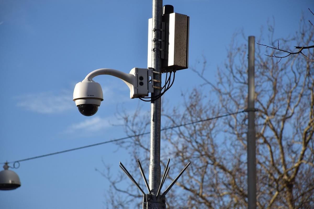 Three white CCTV security cameras mounted together on a vertical metal pole outdoors.