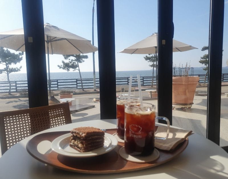 Two iced Americano coffees and a layered chocolate dessert on a table inside a Jebudo ocean view cafe.