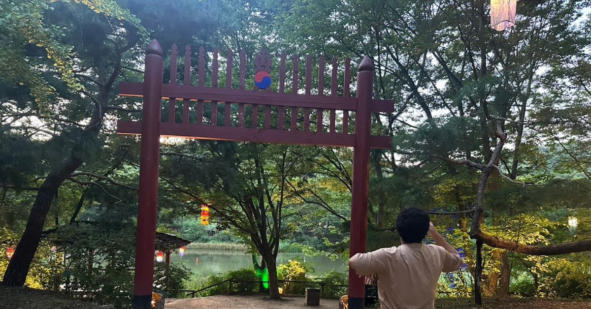 A traditional red wooden gate overlooking a peaceful lake and illuminated trees at dusk in the Korean Folk Village.