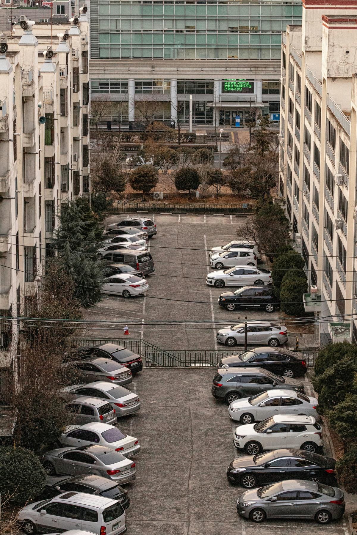 住宅棟の近くにある舗装された屋外駐車場で、数十台の車が非常に狭い間隔で整然と並んで駐車されている様子。