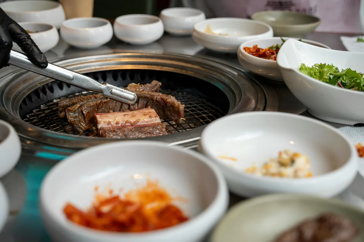 A gloved hand using metal tongs to flip galbi beef on a grill, demonstrating modern korean restaurant etiquette.