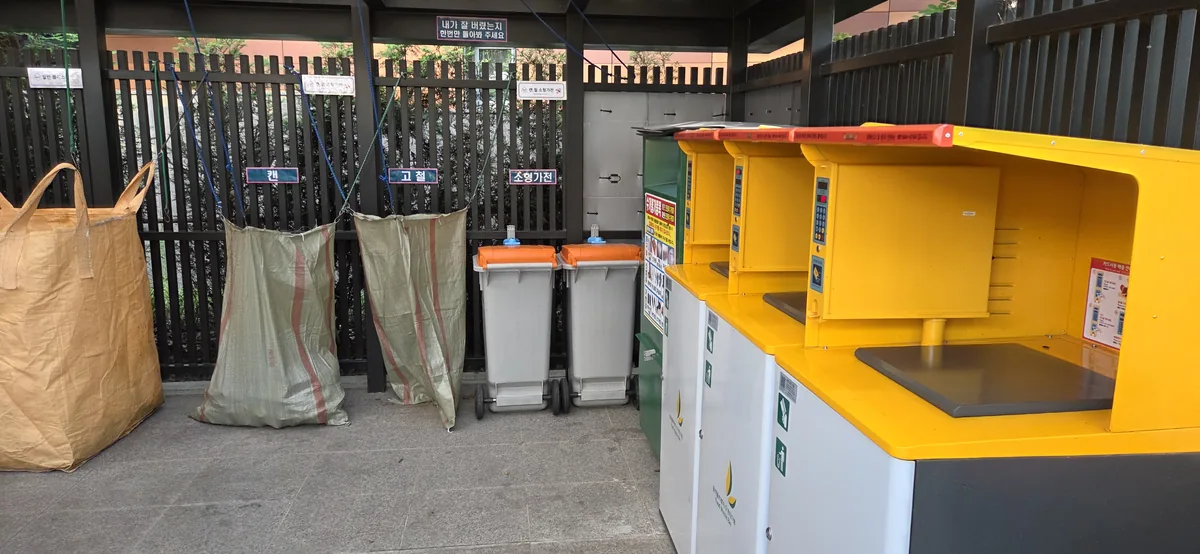 A Korean apartment recycling station with sorting bins, waste bags, and yellow food-waste collection containers.