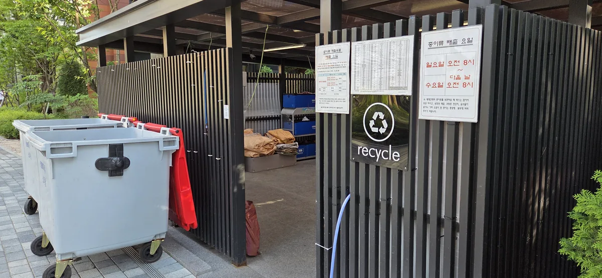 An apartment-complex recycling facility with a white bin, red barrier, Korean signage, and black fencing.