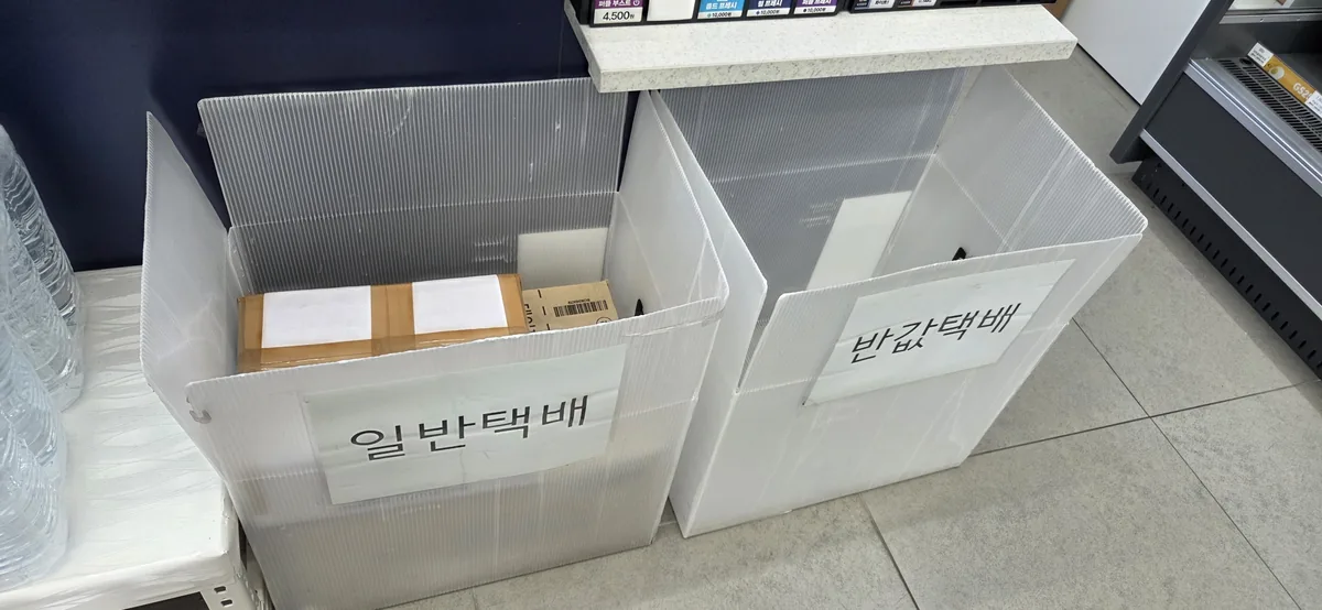 Two white storage bins labeled in Korean sit on a tiled floor at a convenience store counter.