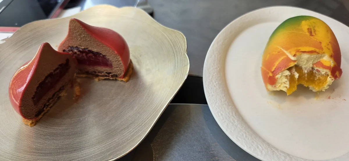 Two plated desserts at a fine dining establishment: a chocolate cake with red glaze on the left, a fruit-topped cake with yellow and orange tones on the right.