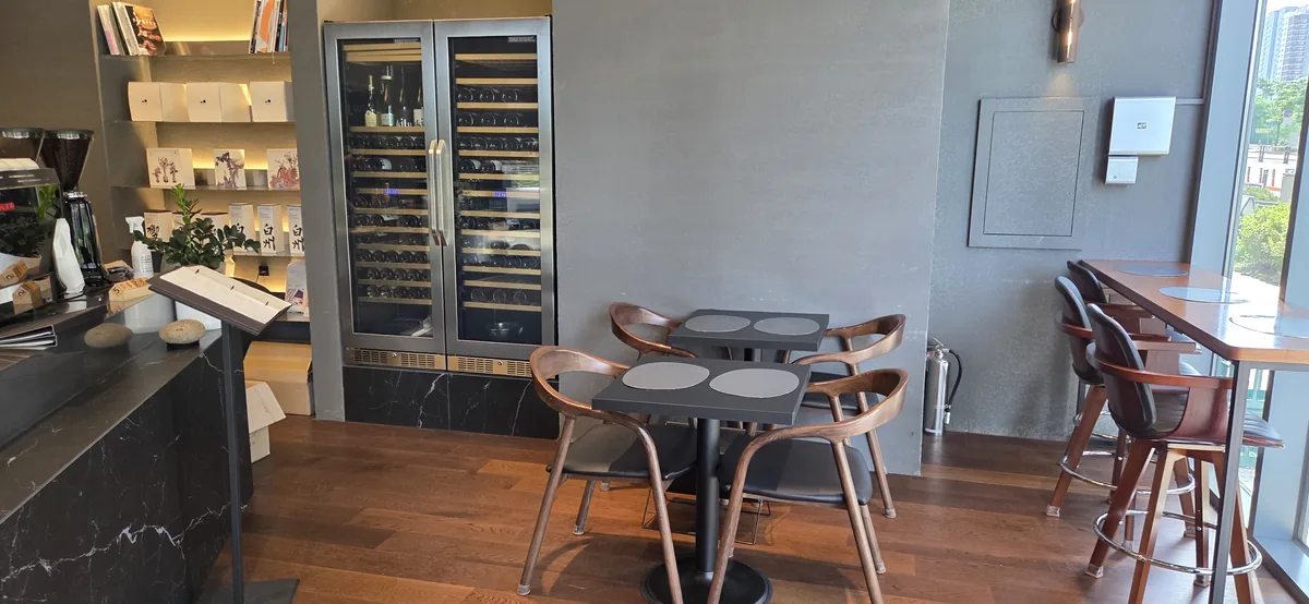 Modern bakery cafe interior with round dining tables, wooden chairs, wine coolers, and shelving behind a service counter.