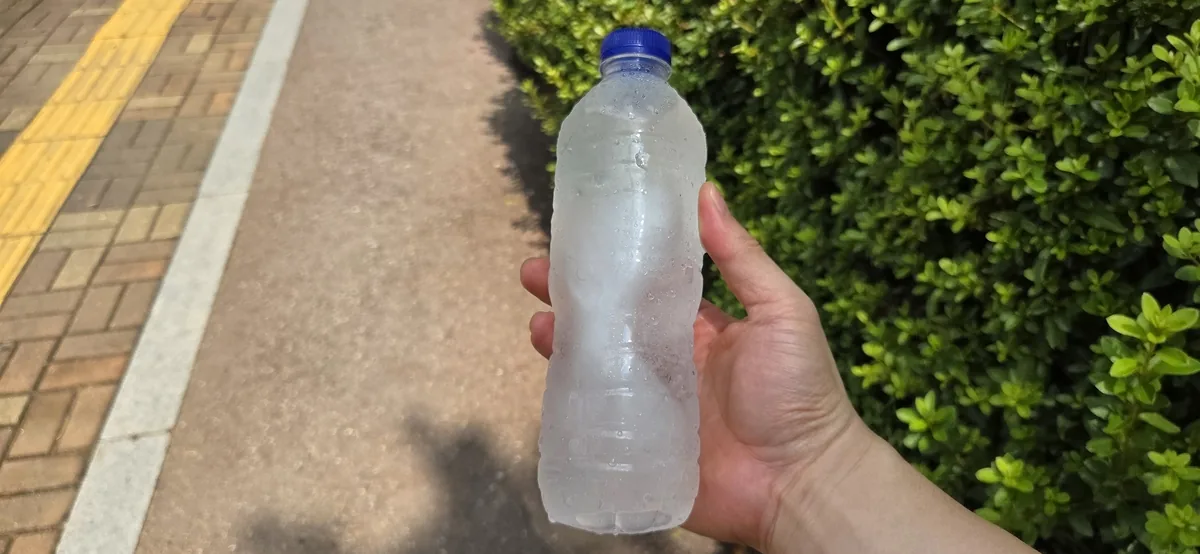 Hand holding a clear plastic bottle with blue cap on a sunny sidewalk.