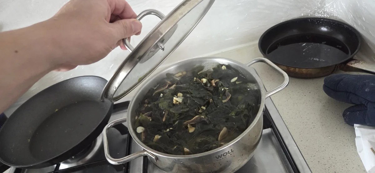 Hand holding a pot lid over a stainless steel pot containing dark seaweed and sliced ingredients on a stovetop.