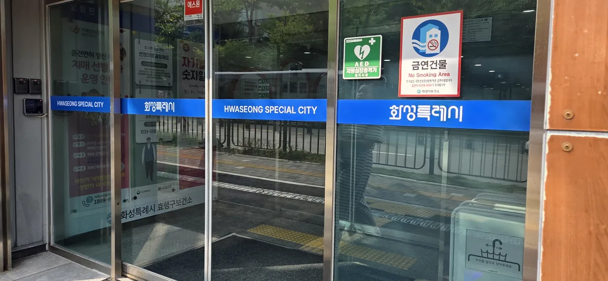 Main entrance doors of the health center, banded in blue with the Hwaseong Special City name in Korean and English.