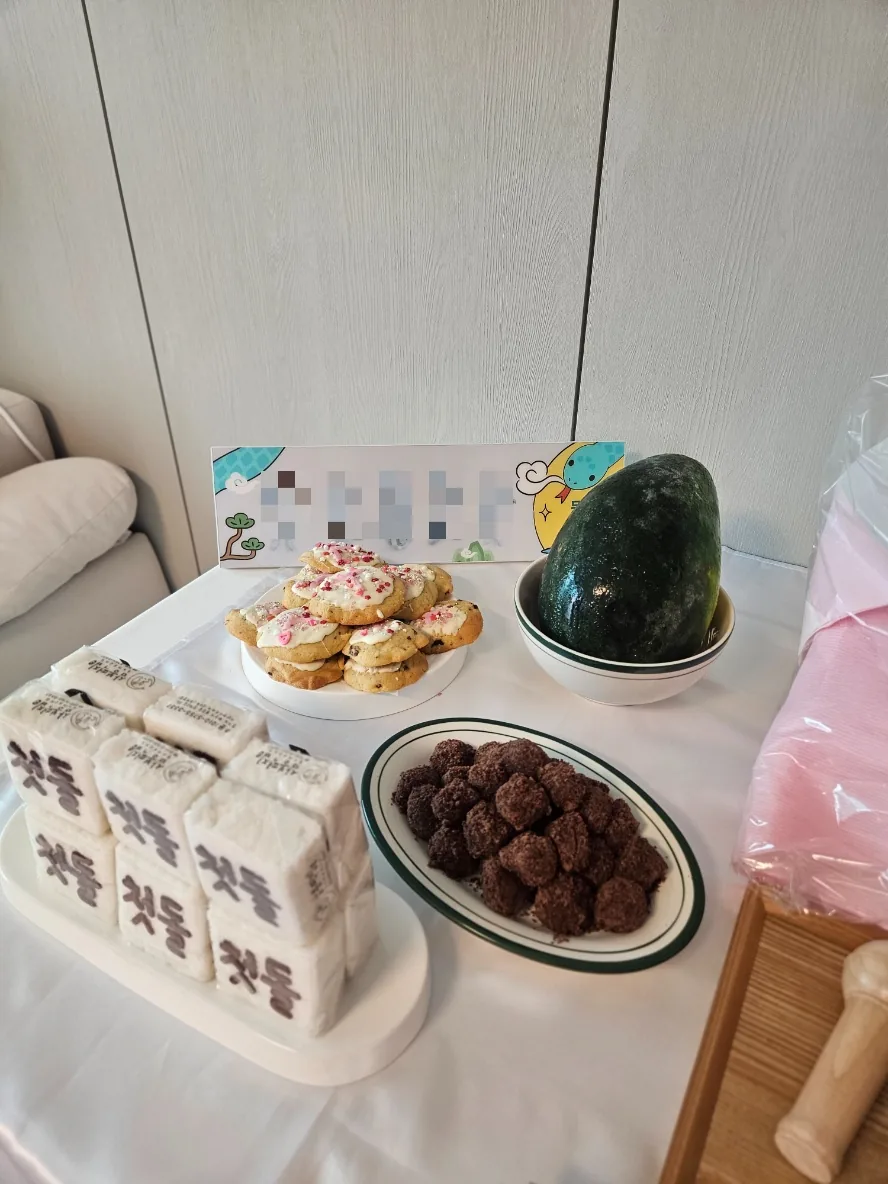 Snack table with wrapped rice cakes stamped 첫돌, iced cookies, chocolate-coated balls and a whole watermelon in a bowl.