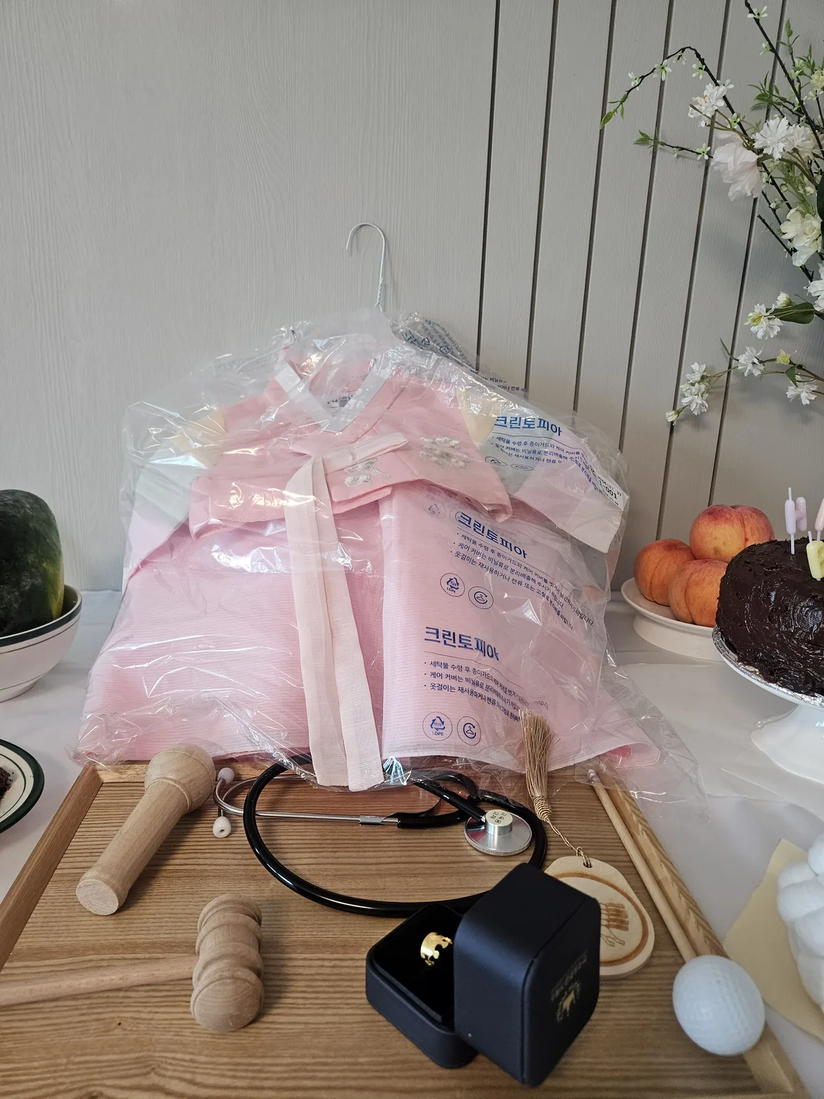 Pink baby hanbok in a dry-cleaning bag on a hanger, with the 돌잡이 items laid out in front: wooden gavel, stethoscope, gold ring in a box, microphone and a golf ball.