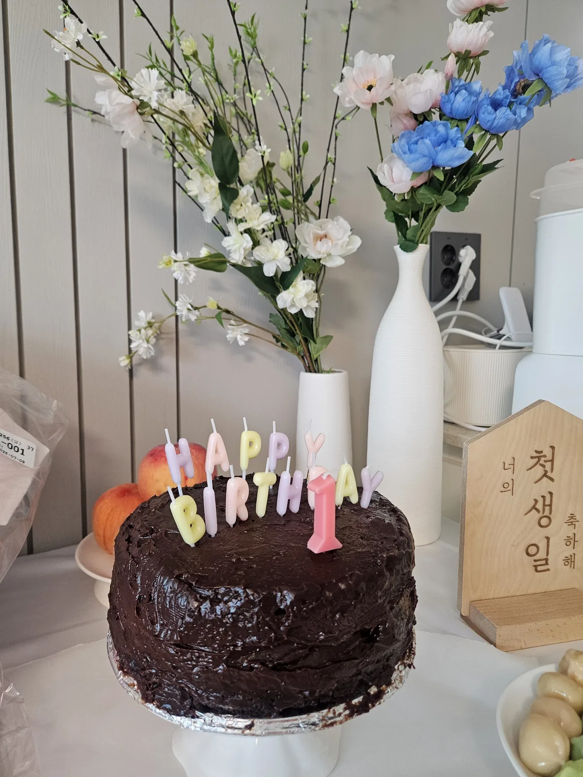 Homemade chocolate cake with pastel HAPPY BIRTHDAY letter candles and a pink number 1, in front of flowers and a wooden sign reading 너의 첫생일 축하해.