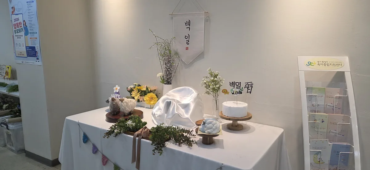 Sample 100-day celebration table displayed at the childcare support center, with a hanging 백일 scroll, flowers, felt sheep, a white cake stand and a 백일 Love cake topper.