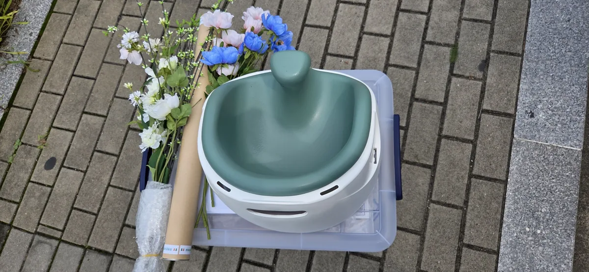 Borrowed baby seat in sage green resting on a clear storage box, next to bundled artificial flowers and a cardboard tube, on a brick pavement outside the center.