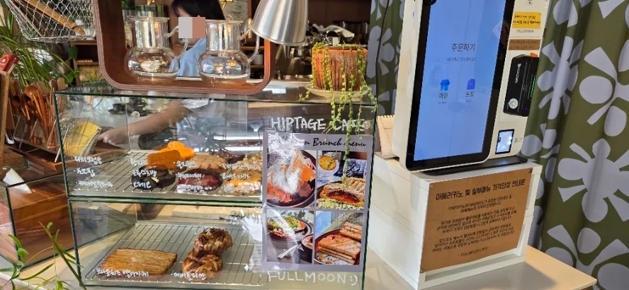 Self-order kiosk at Fullmoon cafe beside a glass case of cookies and pastries with handwritten flavour labels.