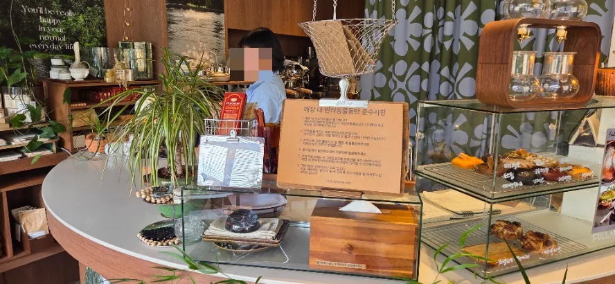 Fullmoon cafe counter with a kraft-paper notice on a clipboard listing the rules for bringing pets inside, next to a pastry case and hanging plants.