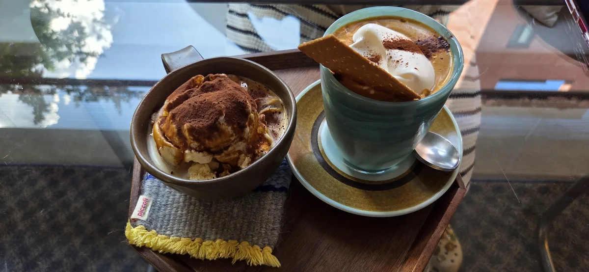 Wooden tray at Fullmoon cafe holding a cream-topped coffee in a pale green glass and a mug of cocoa-dusted dessert on a woven coaster.