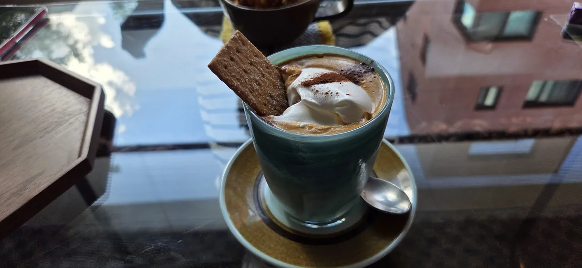 Coffee at Fullmoon cafe in a pale green glass topped with whipped cream, cocoa powder and a biscuit, on a mustard saucer with a spoon.