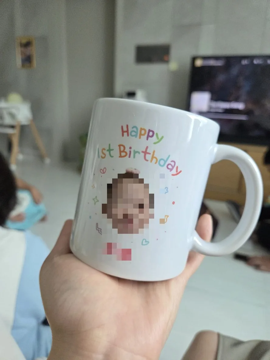 Hand holding a white mug printed Happy 1st Birthday with the baby's photo, the party favor given to each guest.