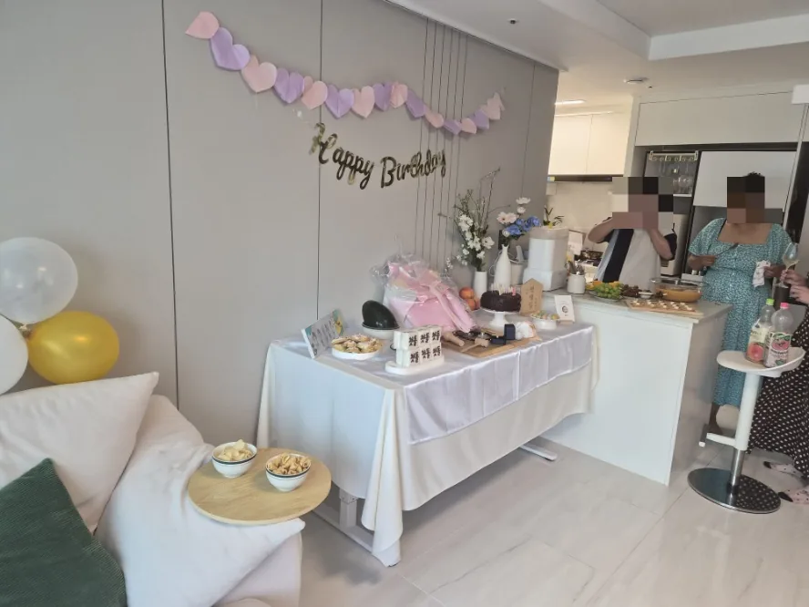 Our living room set up for a home dol party, with a heart garland and Happy Birthday banner over the dol table and guests gathering by the kitchen counter — the answer to how to plan a DIY Doljanchi in Korea on a small budget.