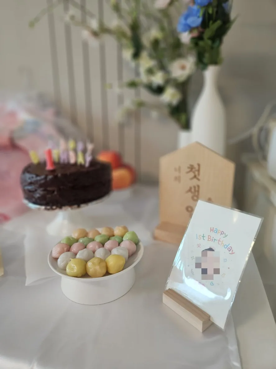 Plate of pastel-colored songpyeon rice cakes beside a small framed Happy 1st Birthday card, with the chocolate cake and 첫생일 sign softly out of focus behind.